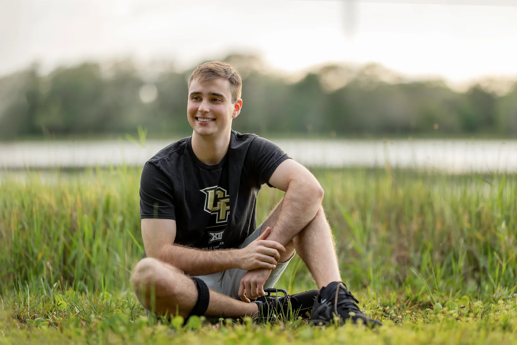 Senior sitting in tall grass beside water at golden hour, relaxed outdoor portrait